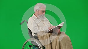 Portrait of aged disabled man on chroma key green screen. Senior man in wheelchair covered in plaid holding reading a