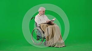 Portrait of aged disabled man on chroma key green screen. Senior man in wheelchair covered in plaid holding reading a