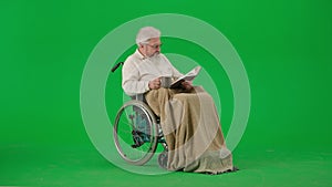 Portrait of aged disabled man on chroma key green screen. Senior man in wheelchair covered in plaid reading book