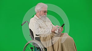 Portrait of aged disabled man on chroma key green screen. Senior man in wheelchair covered in plaid using smartphone.