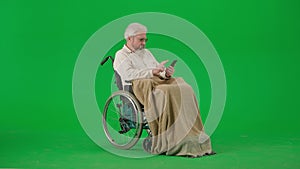 Portrait of aged disabled man on chroma key green screen. Senior man in wheelchair covered in plaid using smartphone.