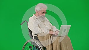 Portrait of aged disabled man on chroma key green screen. Senior man in wheelchair covered in plaid working on laptop.