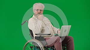 Portrait of aged man on chroma key green screen background. Senior man sitting in wheelchair and holding laptop on knees