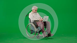 Portrait of aged man on chroma key green screen background. Senior man sitting in wheelchair and holding laptop on knees