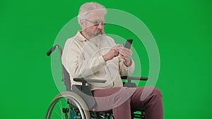 Portrait of aged man on chroma key green screen. Senior man in wheelchair typing bank card number in smartphone, buying