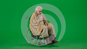 Portrait of aged man isolated on chroma key green screen background. Senior man in wheelchair covered by plaid drinking