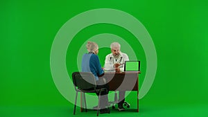 Portrait of aged man medic on chroma key green screen. Senior doctor at table talking with woman patient, showing on