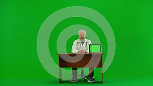 Portrait of aged man medic on chroma key green screen. Senior doctor in uniform sitting at table showing information on