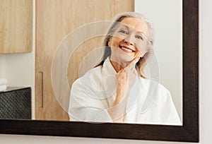 Portrait of an aged woman in a bathroom. Smiling woman with grey hair looking at her reflection in a mirror