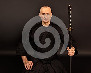 Portrait of aikido sensei master with black sensei belt in taekwondo kimono witn sword katana on black background. Traditional