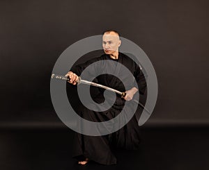 Portrait of aikido sensei master with black sensei belt in taekwondo kimono witn sword katana on black background. Traditional