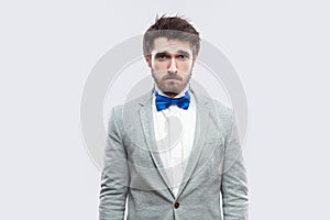 Portrait of alone handsome bearded man in casual grey suit and blue bow tie standing and looking at camera with sad eyes