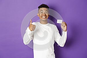 Portrait of amazed african-american man showing thumbs-up and credit card, recommend bank loan, standing over purple