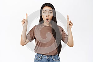 Portrait of amazed asian girl points at banner, shows loog with surprised, impressed face expression, stands over white