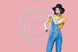 Portrait of amazed beautiful young woman in yellow t-shirt and blue denim overalls with makeup and black hat standing and pointing