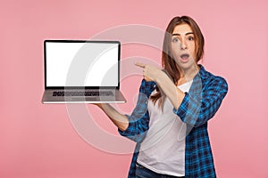 Portrait of amazed charming girl in checkered shirt showing blank laptop screen with shocked expression