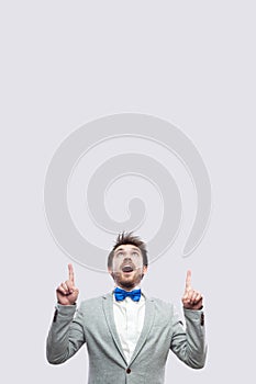 Portrait of amazed handsome bearded man in casual grey suit and blue bow tie standing looking and pointing at up copyspace