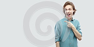 Portrait of amazed handsome long haired blonde young man in blue casual shirt standing and pointing at side copyspace with