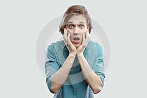 Portrait of amazed handsome long haired blonde young man in blue shirt standing with big eyes holding hand on face and looking