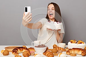 Portrait of amazed happy beautiful woman with brown hair wearing white T-shirt sitting at table isolated over gray background,