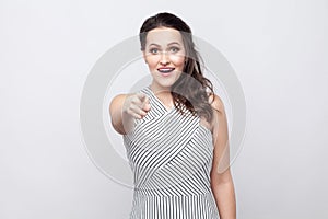 Portrait of amazed happy beautiful young brunette woman with makeup and striped dress standing, looking and pointing at camera