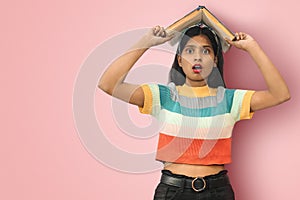 Portrait of an amazed indian asian girl student with open mouth and big eyes posing  holding book on her head and looking
