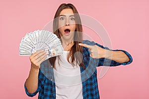 Portrait of amazed lucky girl in casual shirt excited and surprised to hold lot of money, pointing at dollar bills