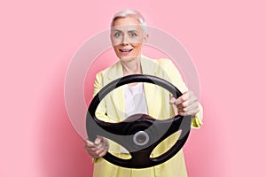 Portrait of amazed mature age funny business lady wear yellow suit driving her husband automobile isolated over pink