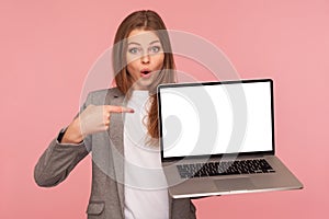 Portrait of amazed pretty brunette businesswoman in elegant suit pointing at mock up display, holding laptop