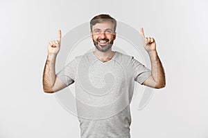 Portrait of amazed smiling guy with a beard, wearing grey t-shirt, pointing fingers up at logo, standing over white
