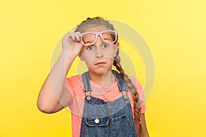 Portrait of amazed stylish little girl in denim overalls taking off pink eyeglasses and looking with astonished expression