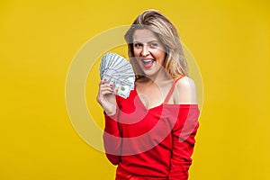 Portrait of amazed wealthy young woman in red dress holding money, isolated on yellow background