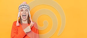 Portrait of amazed woman wearing orange jumper and knitted earflap hat standing isolated over yellow background, pointing away,