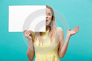 Portrait of amazed young blond woman holding blank sign with copy space on blue studio background. Showing shocked