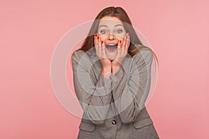 Portrait of amazed young brunette woman in business suit keeping hands on face and shouting in surprise