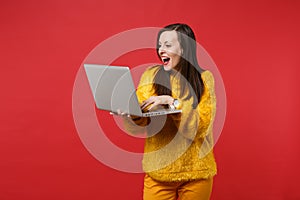 Portrait of amazed young woman in yellow fur sweater keeping mouth wide open using laptop pc computer isolated on bright