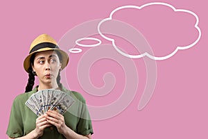 Portrait of a amazement Caucasian woman wearing straw hat and holding a fan of dollars banknotes. Pink background with