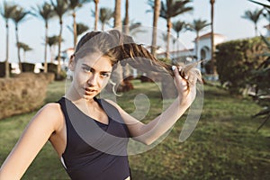 Portrait amazing attractive young woman in sportswear playing with her long curly hair in park of tropical city. Looking