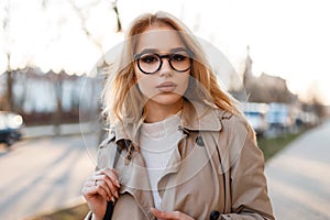 Portrait of amazing cute young hipster woman with beautiful lips in stylish glasses in a spring stylish coat in a white T-shirt