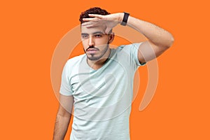 Portrait of ambitious confident man holding hand over eyes and looking far. indoor studio shot isolated on orange background