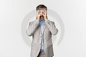 Portrait of ambushed businessman in grey suit, cover eyes and peeking through fingers with scared face, standing over