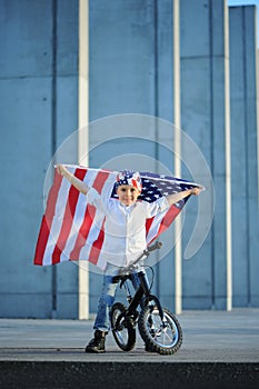 A portrait of american boy sitting on bike waving american flag.