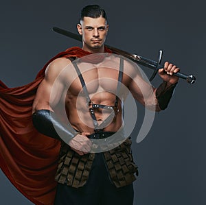 Portrait of ancient Greece warrior with a muscular body dressed in battle uniforms holds sword on his shoulder.