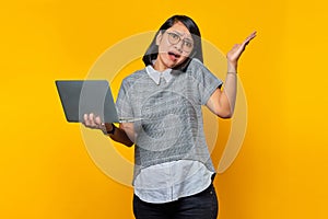Portrait of angry asian woman talking on smartphone while holding laptop on yellow background