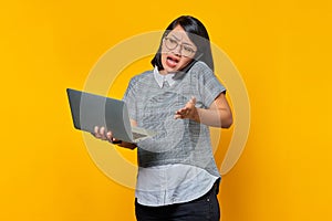 Portrait of angry asian woman talking on smartphone while holding laptop on yellow background