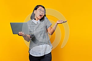 Portrait of angry asian woman talking on smartphone while holding laptop on yellow background