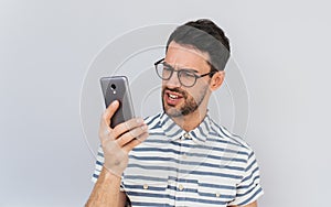 Portrait of angry bearded handsome businessman wears round spectacles and casual shirt, looks furiously into mobile phone, posing