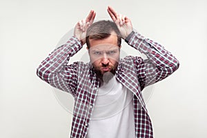 Portrait of angry bearded man showing bull horn gesture with puffed out cheeks to look more menacing. white background
