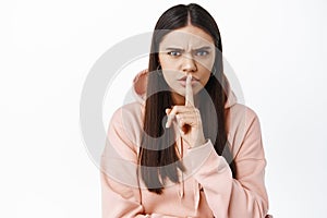 Portrait of angry and bossy young woman frowning, shushing at camera with shh taboo sign on lips, tell to keep quiet