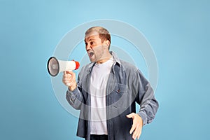 Portrait of angry crazy wild young man shouting to loudspeaker isolated blue studio background. Irritated young guy in